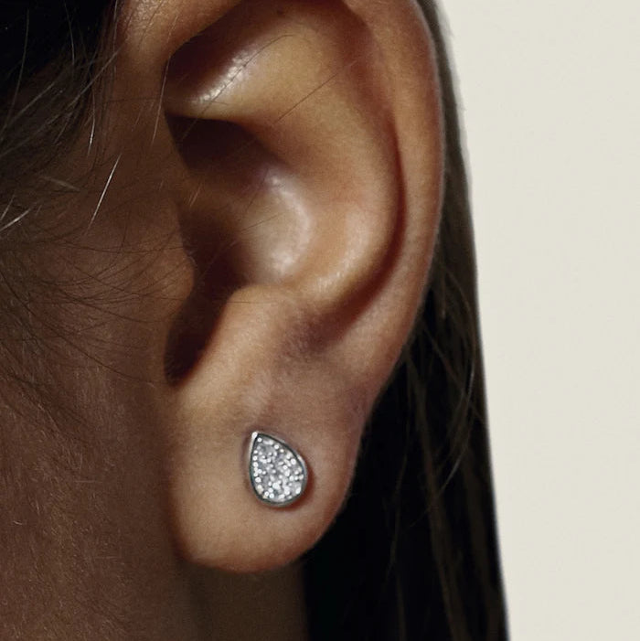 A close-up of a person's ear wearing a small teardrop-shaped stud earring with a clear crystal-like center and a silver-toned border.