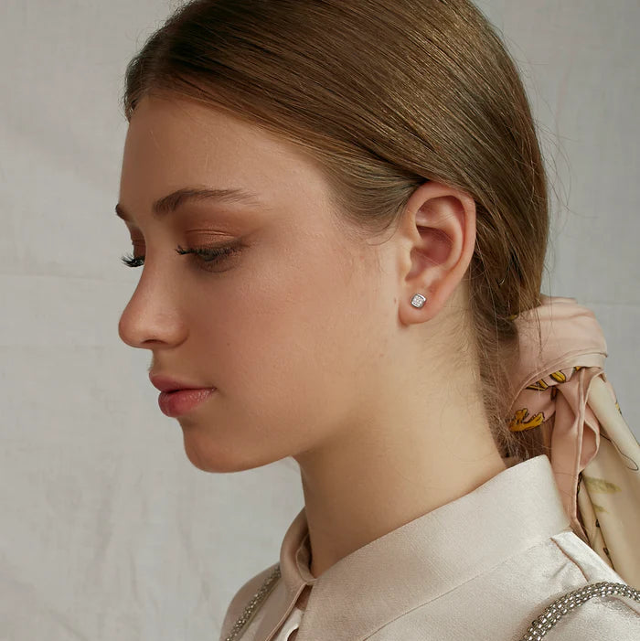 A close-up side profile of a woman with light brown hair tied back, wearing a pale blush satin shirt with silver beaded trim and a matching satin hair tie with a small gold floral accent. She is wearing a small silver stud earring.