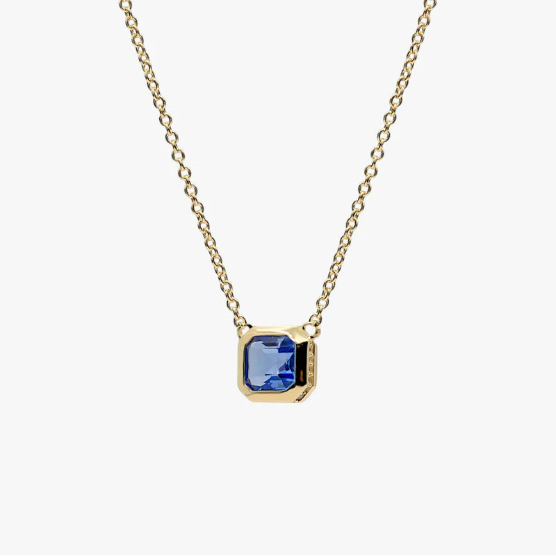 A delicate gold chain necklace featuring a small square gold pendant with a blue gemstone, set against a plain light gray background.