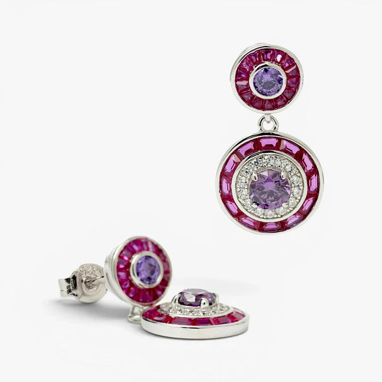 A pair of silver-tone drop earrings featuring circular designs with a purple gemstone in the center, surrounded by clear crystals and a halo of pink gemstones, set against a plain white background.