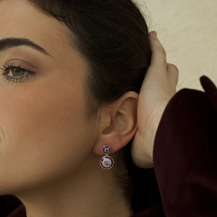 A close-up of a woman wearing a small silver-toned dangle earring with a purple top stone and a circular clear stone drop, paired with a dark burgundy velvet jacket.