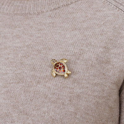 A small gold turtle-shaped brooch with red and white enamel detailing, pinned to the chest of a beige knit sweater.