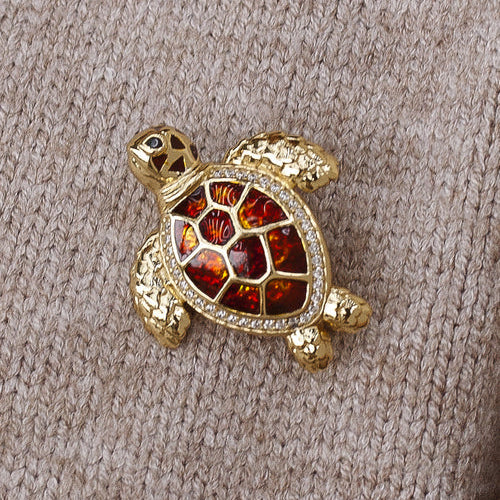 A gold turtle-shaped brooch with a red and orange enamel shell, set against a beige knit fabric background.