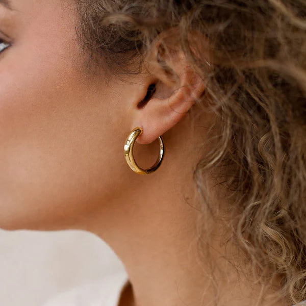 A close-up side profile of a woman wearing a small gold hoop earring, with curly light brown hair and a white t-shirt against a soft neutral background.