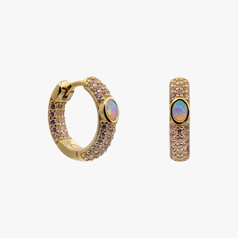 A pair of gold huggie hoop earrings decorated with rows of small pinkish-purple crystals and a central iridescent opal-like stone, shown on a plain light gray background.