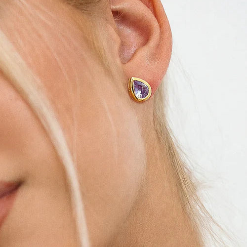 A close-up of a model's ear wearing a gold teardrop stud earring with a purple amethyst gemstone, set against a clean white background.