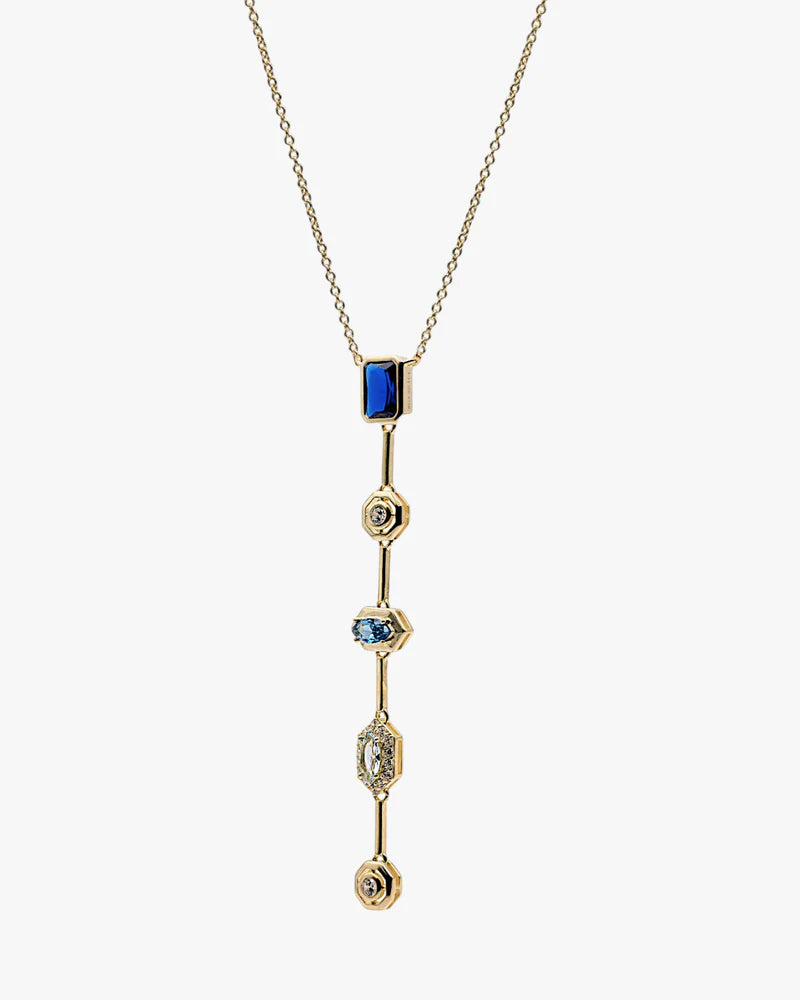 A gold chain necklace with a long, vertical drop pendant featuring a rectangular blue stone at the top and three smaller blue and clear gemstone accents on a light gray background.