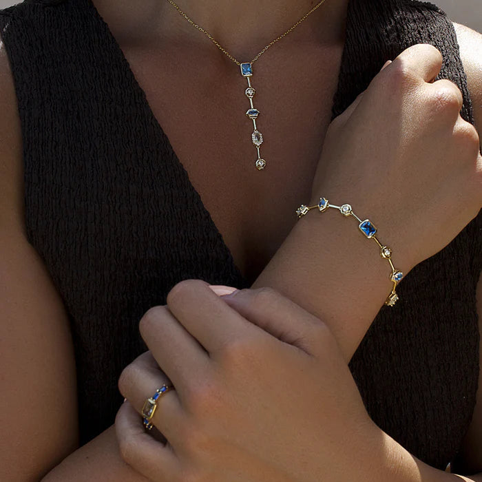A close-up lifestyle shot of a model wearing a delicate gold necklace with a vertical drop pendant and a matching gold bracelet, both featuring small blue and clear gemstones.