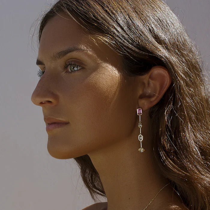A close-up side profile of a woman wearing a delicate gold chain necklace and matching drop earrings with pink gemstone accents, set against a neutral light gray background.