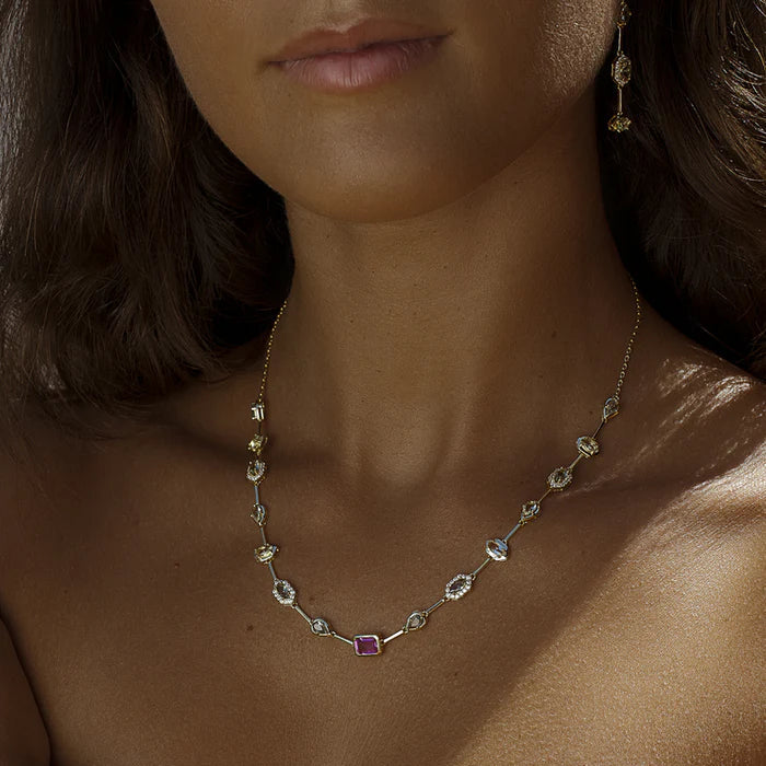 A close-up of a model wearing a delicate gold necklace with small clear stones and a central pink gemstone, paired with matching gold drop earrings and a bracelet.