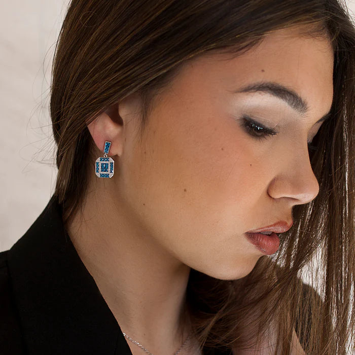 A close-up of a woman wearing a silver-toned necklace and matching drop earring, both featuring a rectangular blue gemstone pendant, against a light neutral background.
