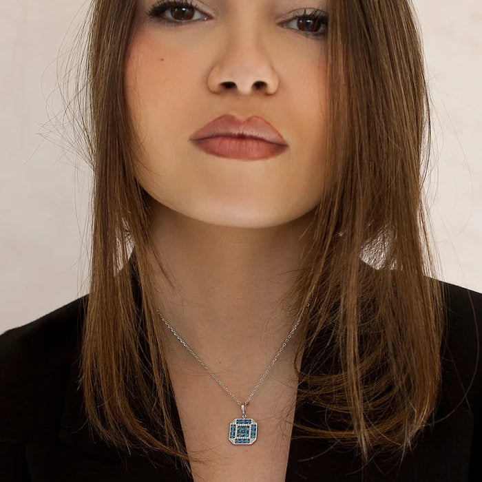 A close-up of a woman with brown hair wearing a silver chain necklace with a square pendant featuring a blue and white geometric pattern, set against a plain light background.