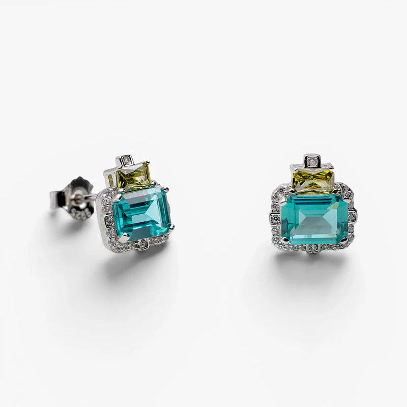 A pair of silver stud earrings featuring a large emerald-cut turquoise blue gemstone and a smaller yellow-green gemstone, accented with small clear stones, shown on a white background.