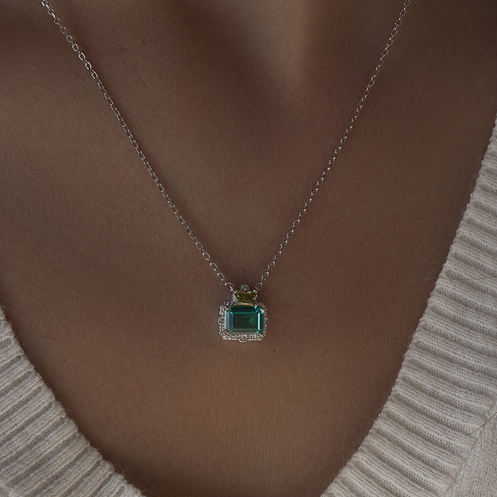 A close-up of a person wearing a delicate silver chain necklace with a square emerald-cut green gemstone pendant, styled with a ribbed cream-colored V-neck sweater.