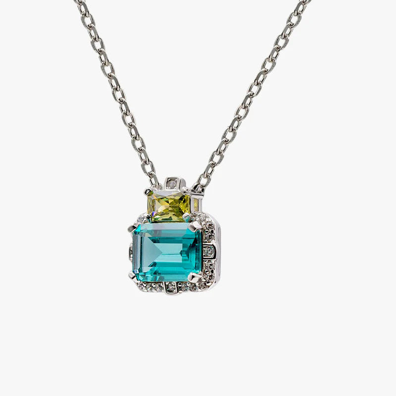 A silver-toned necklace featuring a pendant with a large emerald-cut turquoise-blue gemstone and a smaller yellow-green gemstone, set against a plain light gray background.