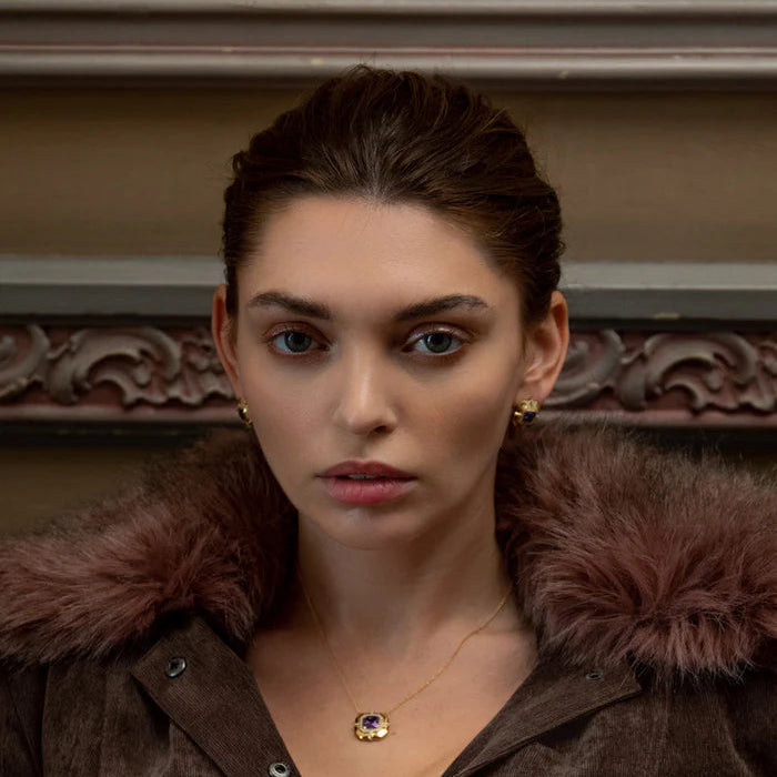 A close-up portrait of a woman wearing a brown suede jacket with a dark brown fur collar, a delicate gold necklace with a round purple and gold pendant, and small gold earrings, set against an ornate brown background.